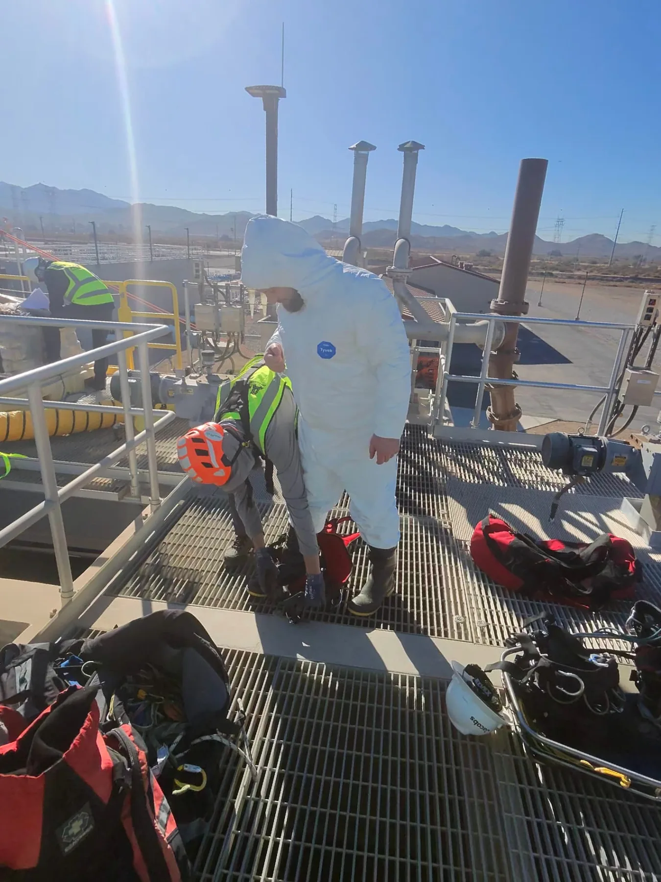 Worker in a protective coverall being assisted with equipment on an industrial platform