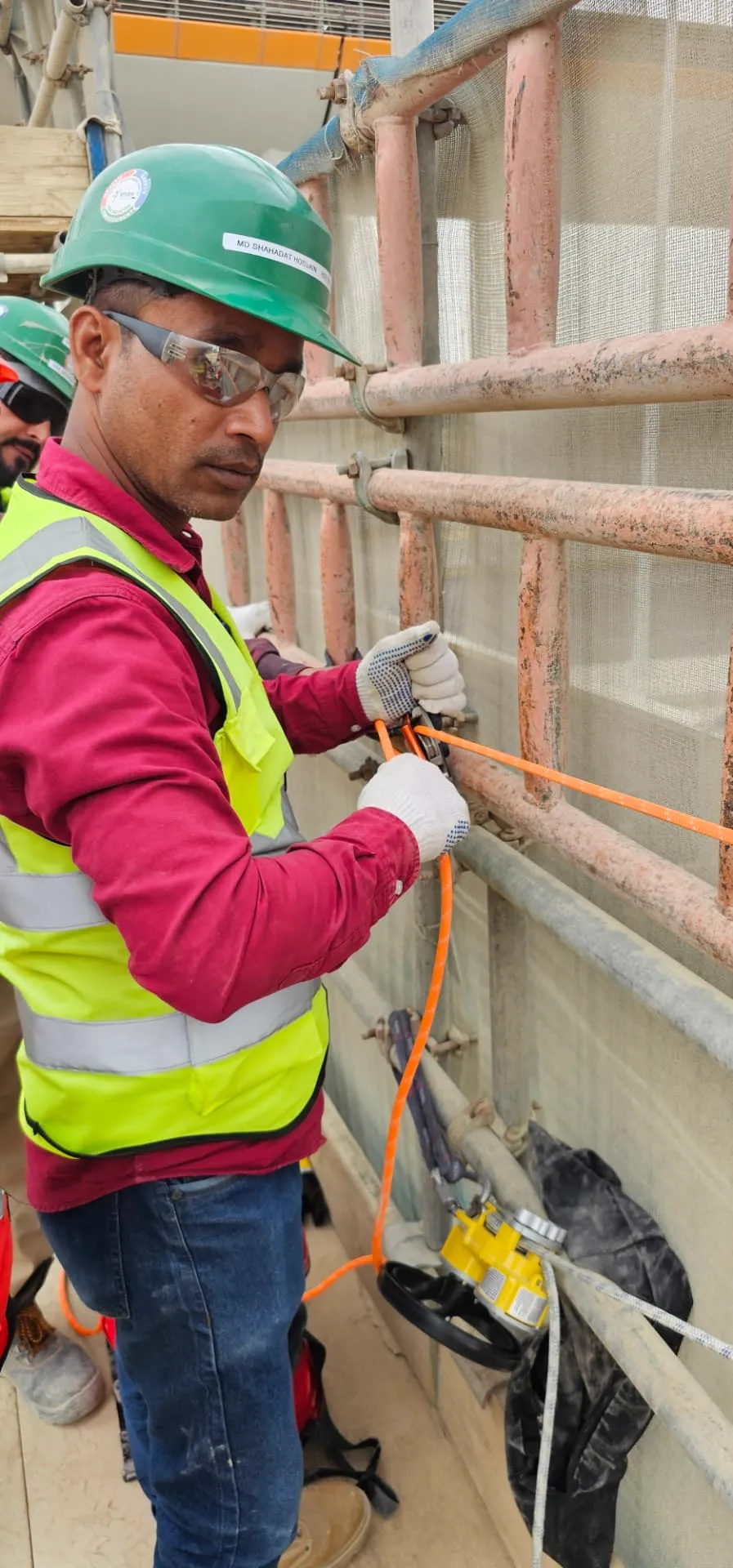 Technician in high-visibility PPE connecting an orange lifeline to a scaffold anchor point