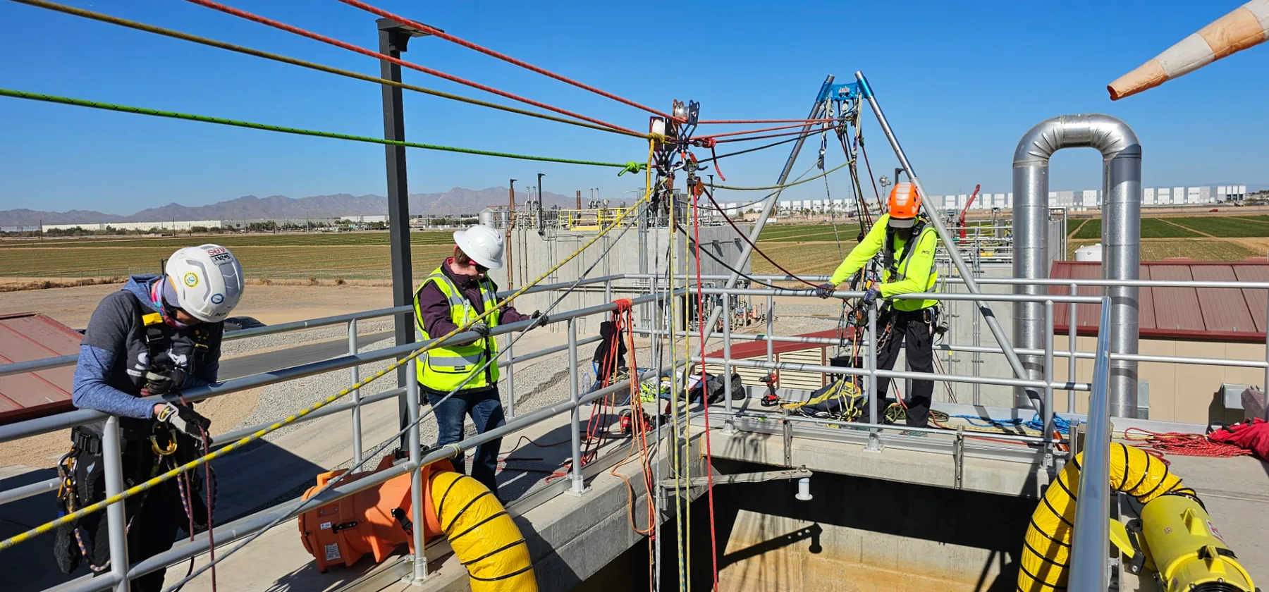 Three technicians managing color-coded rope systems rigged over an open industrial vault